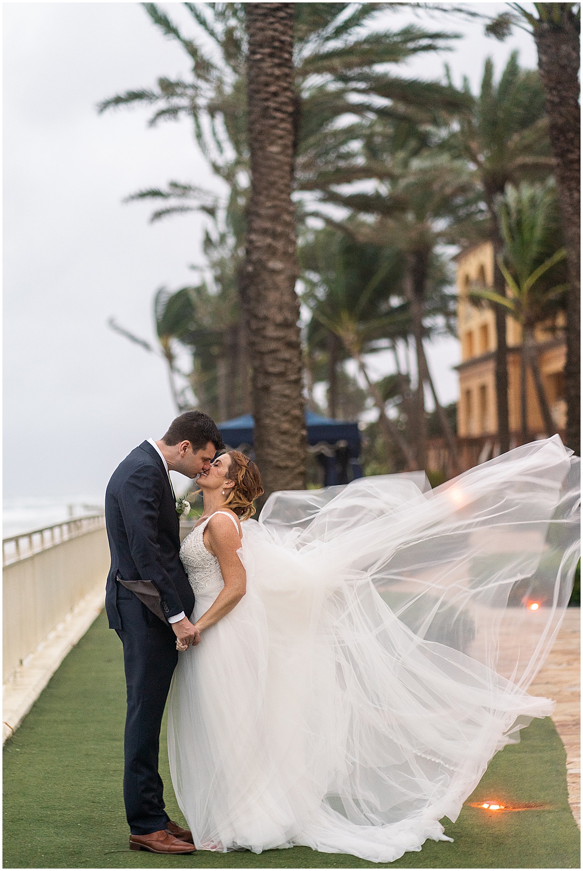 Bride and groom kissing beneath palm trees as the bride’s veil blows in the wind at a Palm Beach wedding.