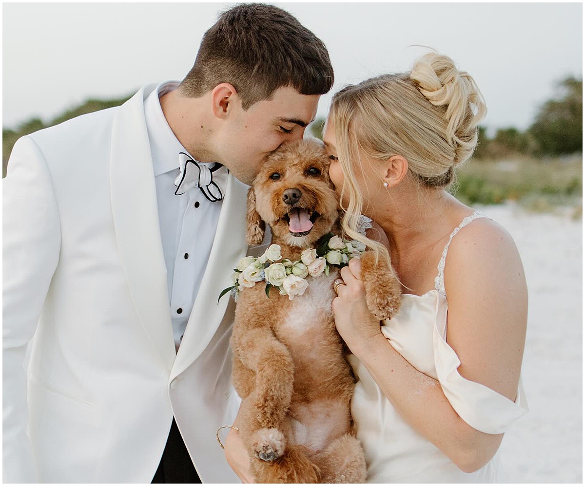 Bride and groom kiss their smiling dog during their wedding portraits